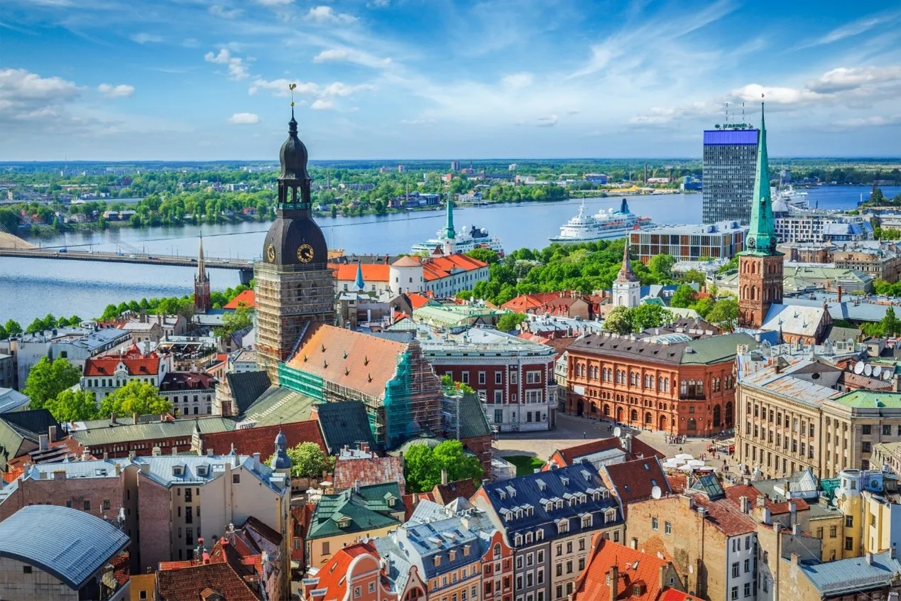 Panoramic view of Riga’s Old Town with historic red-roofed buildings and church towers, including St. Peter’s Church in the foreground, overlooking the Daugava River and a modern bridge under a blue sky.
