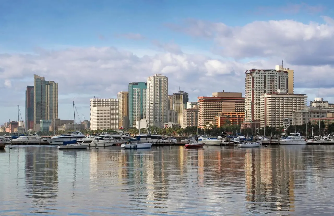 Manila skyline with high-rise buildings and a marina filled with boats reflected in calm waterfront waters.