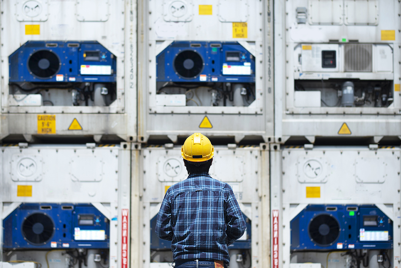 Worker inspecting stacked refrigerated shipping containers, ensuring safety compliance and operational readiness