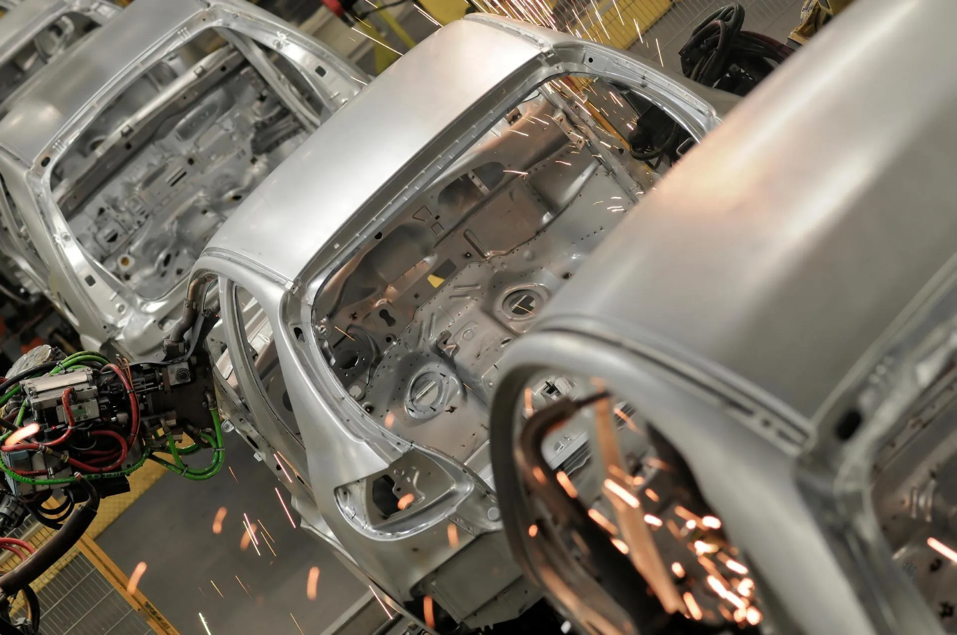 Automated robotic arms weld silver car body frames on an assembly line, producing bright sparks inside a modern automotive factory.