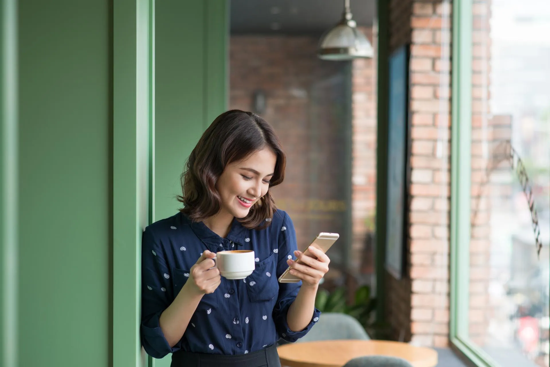 A woman in a blue shirt reads a message on her phone while holding a coffee cup.
