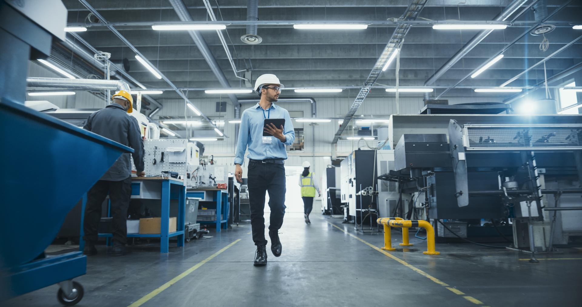 A worker wearing a white hard hat carries a tablet while walking between production equipment and automated machines at an industrial manufacturing facility.