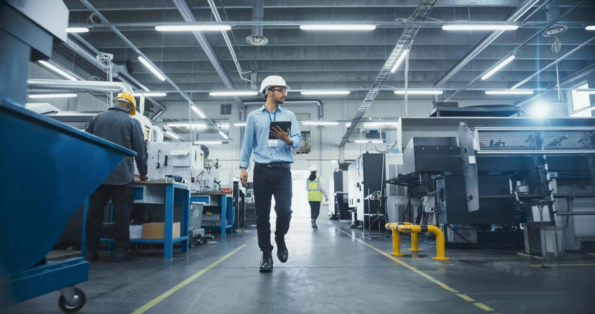 A worker wearing a white hard hat carries a tablet while walking between production equipment and automated machines at an industrial manufacturing facility.