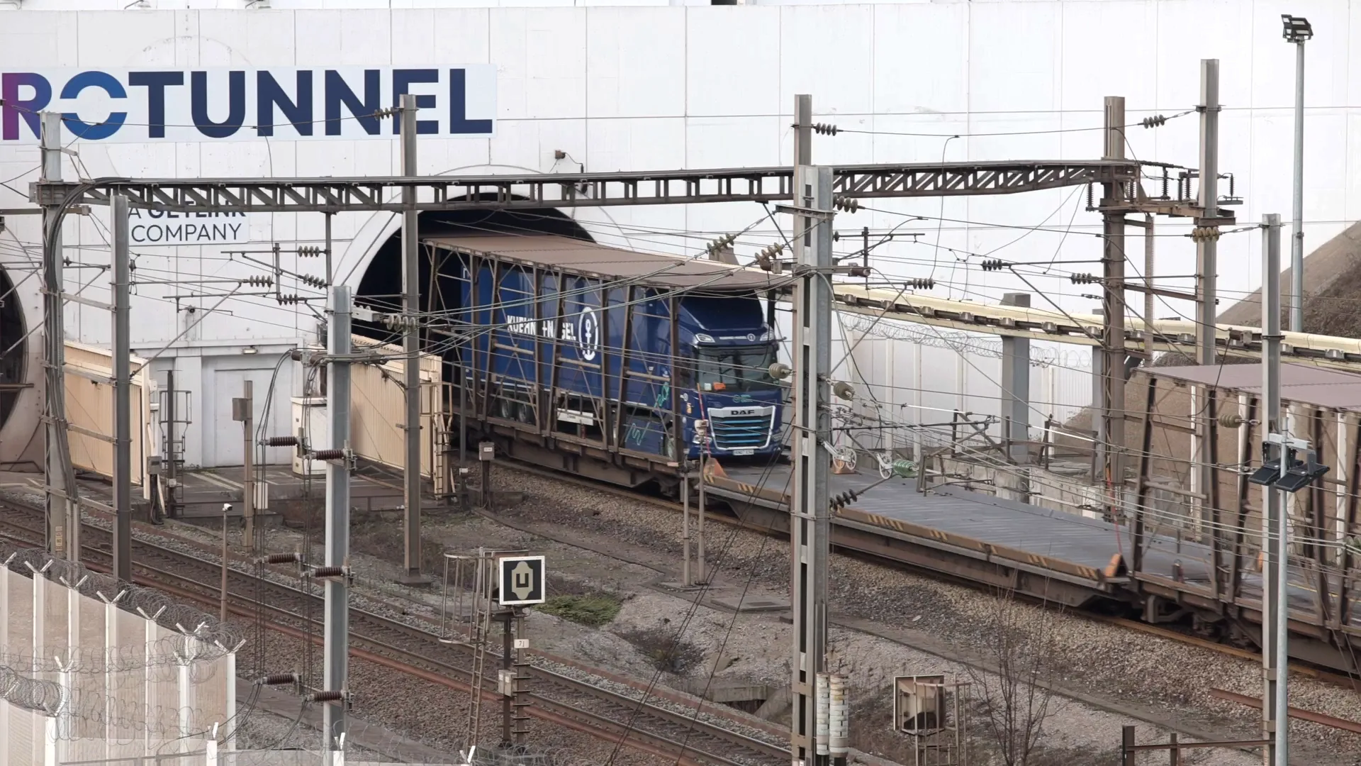 Blue Kuehne+Nagel electric DAF truck exiting a Eurotunnel freight shuttle platform surrounded by railway infrastructure.