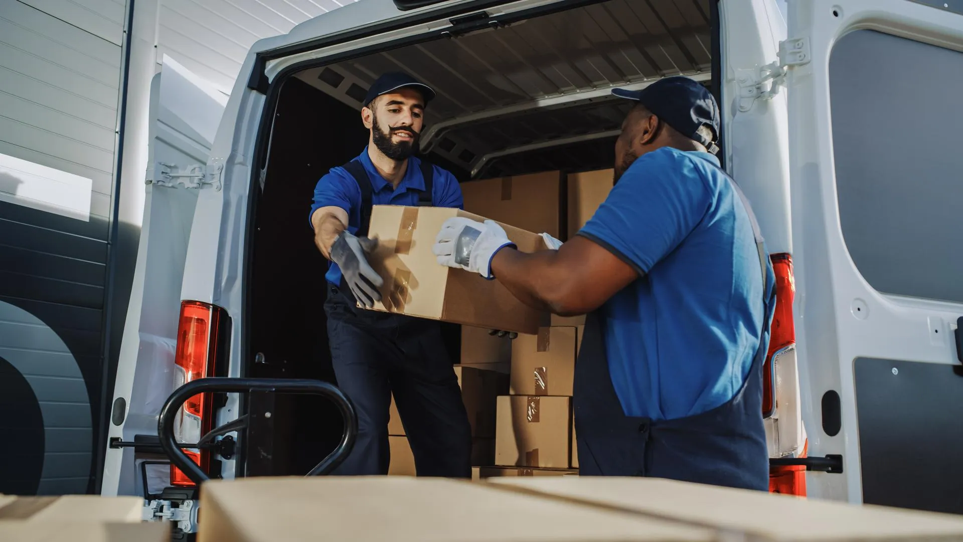 Logistics Warehouse Two Happy Workers Talk, Joke use Hand Pallet Truck Start Loading Delivery Truck with Cardboard Boxes, Online Orders, Purchases, E-Commerce Goods.