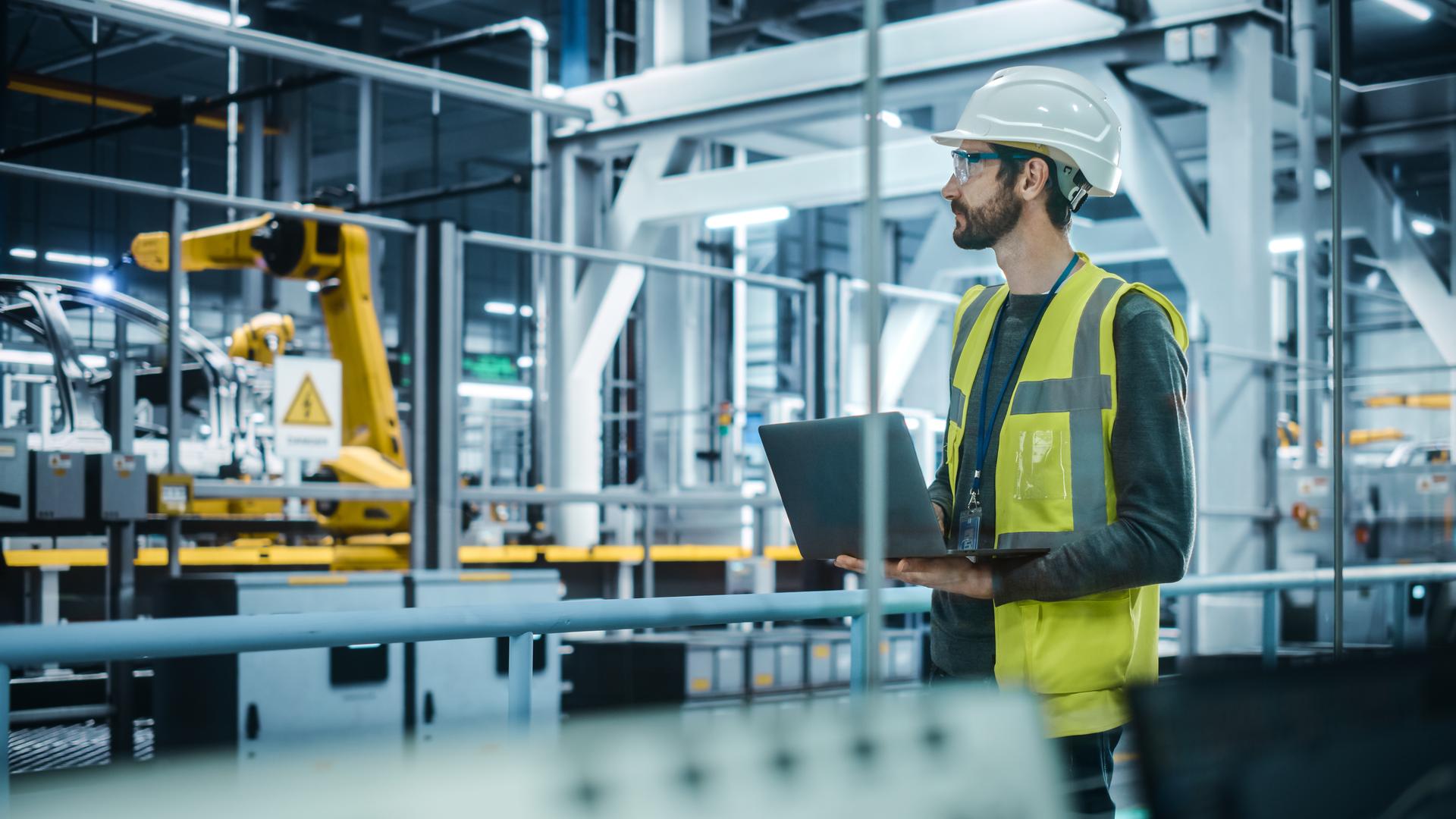 Car Factory: Male Automotive Engineer Wearing Hard Hat, Standing, Using Laptop. Monitoring, Control, Equipment Production. Automated Robot Arm Assembly Line Manufacturing Electric Vehicles