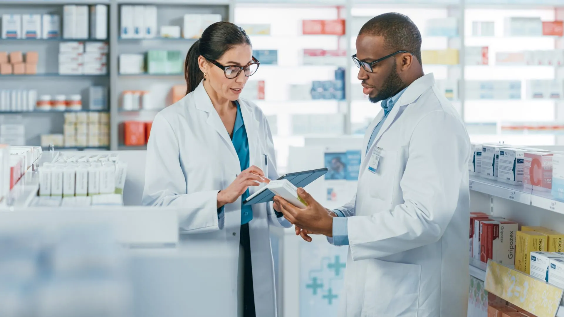 Two pharmacy professionals reviewing medication packaging and digital inventory information in a retail pharmacy.