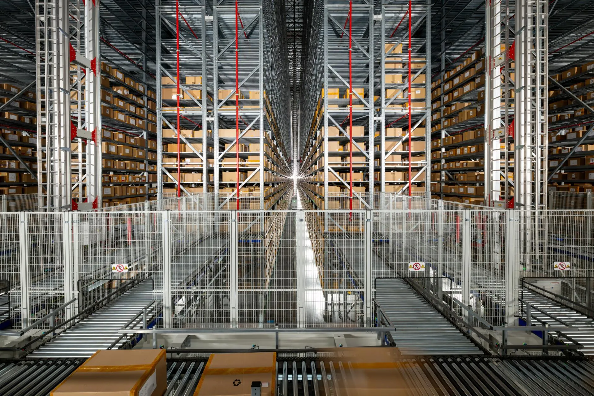 Automated warehouse storage system with tall metal shelving racks extending into the distance and filled with neatly organized cardboard boxes. Conveyor rollers and safety fencing are visible in the foreground.