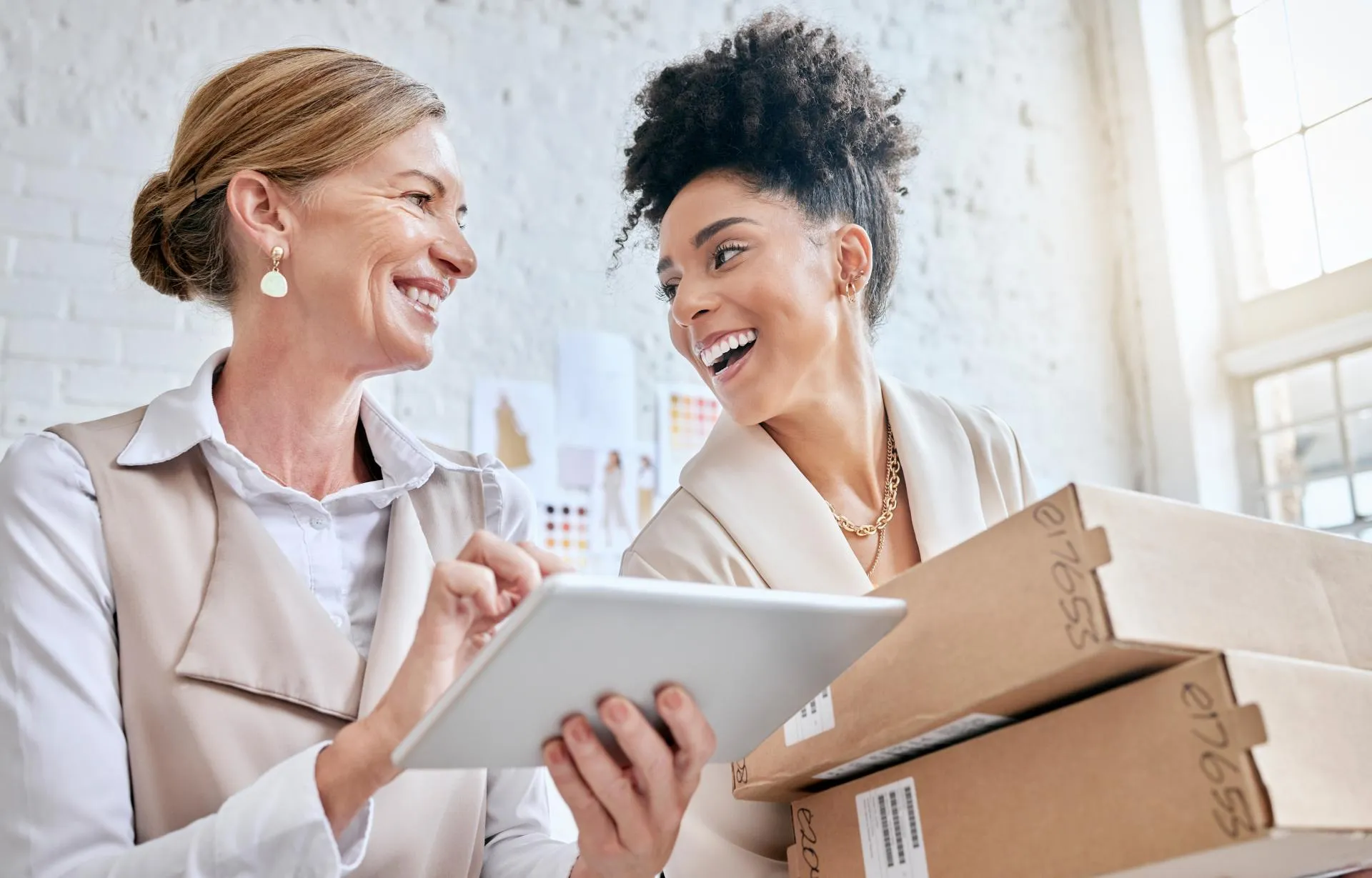 Two retail employees in a bright workspace smile and review information on a tablet while handling stacked shoe boxes.