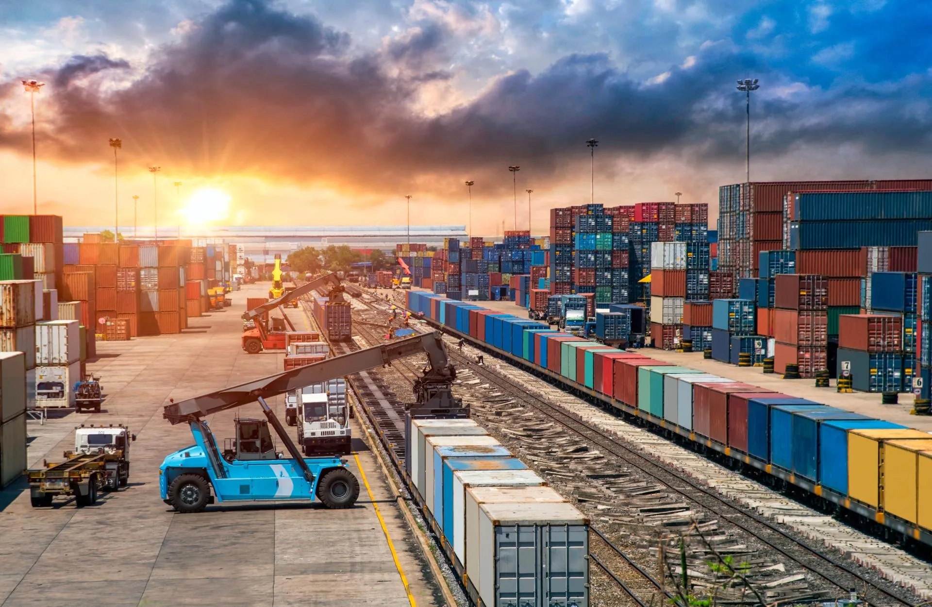 Containers stacked along tracks with vehicles loading and a sunset in the background