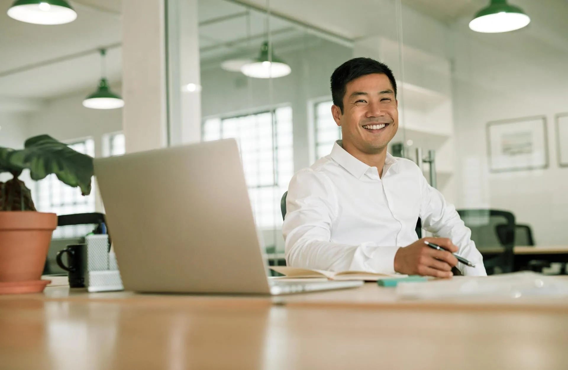 Professional smiling and sitting at a desk with a laptop and notebook in a modern office environment.