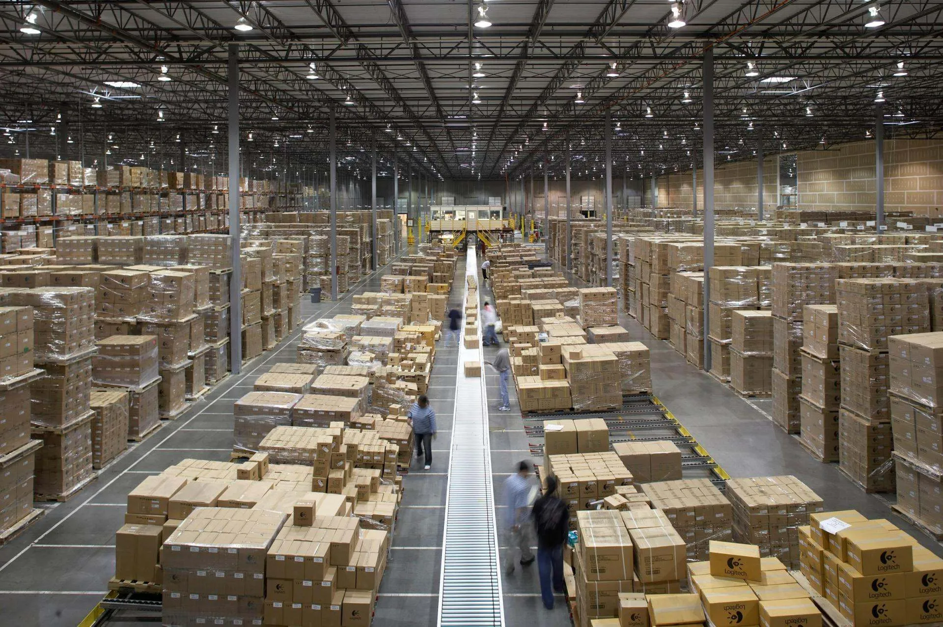 Large logistics warehouse with organized pallets, workers operating along a conveyor belt, showcasing efficient inventory and distribution processes.