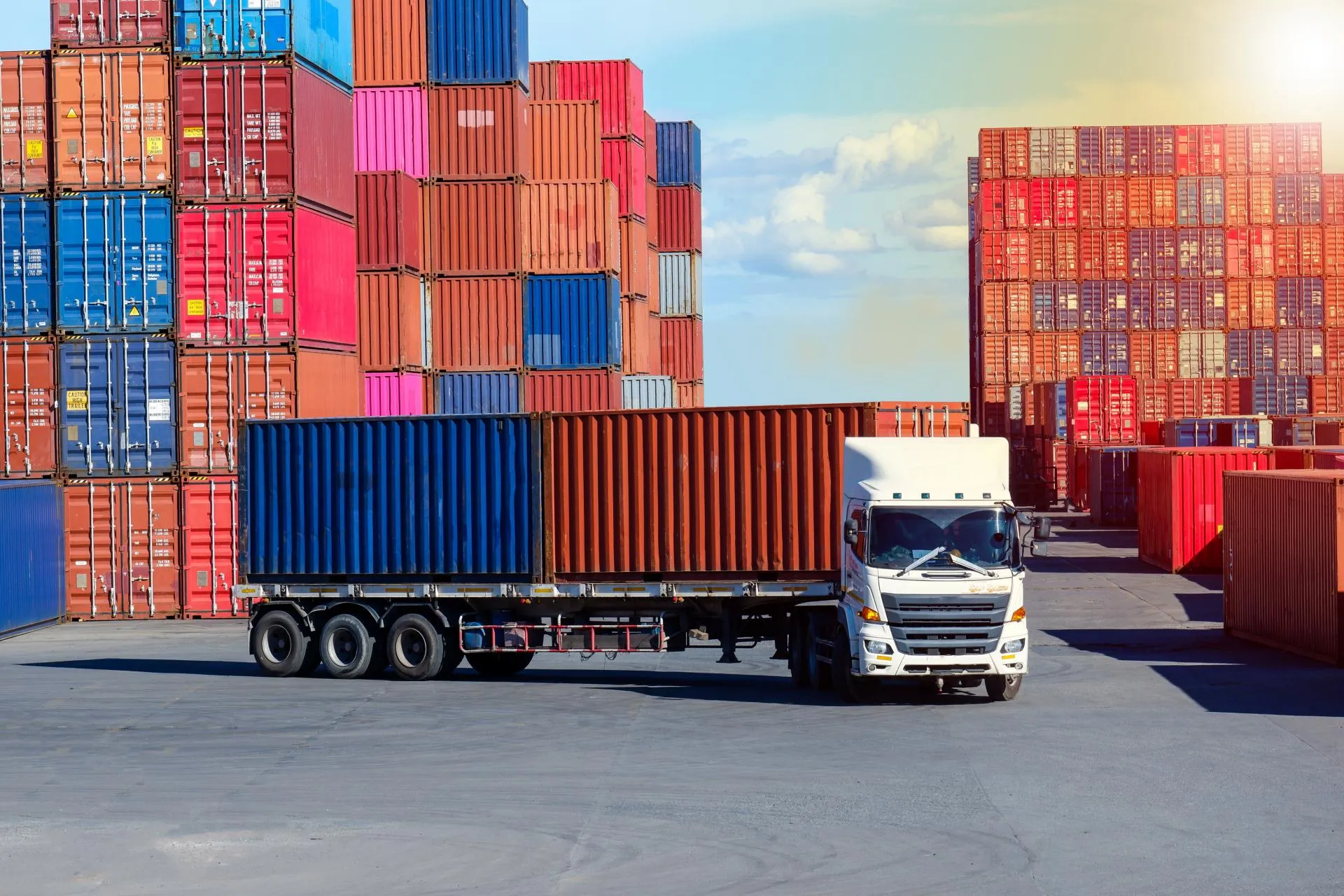 A white truck moves past stacked red, blue, pink, and orange shipping containers on a paved lot.