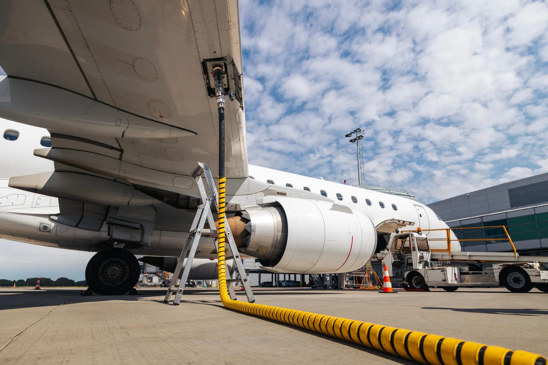 Aircraft being refueled on the airport apron, with a fuel hose connected under the wing and ground service equipment nearby.