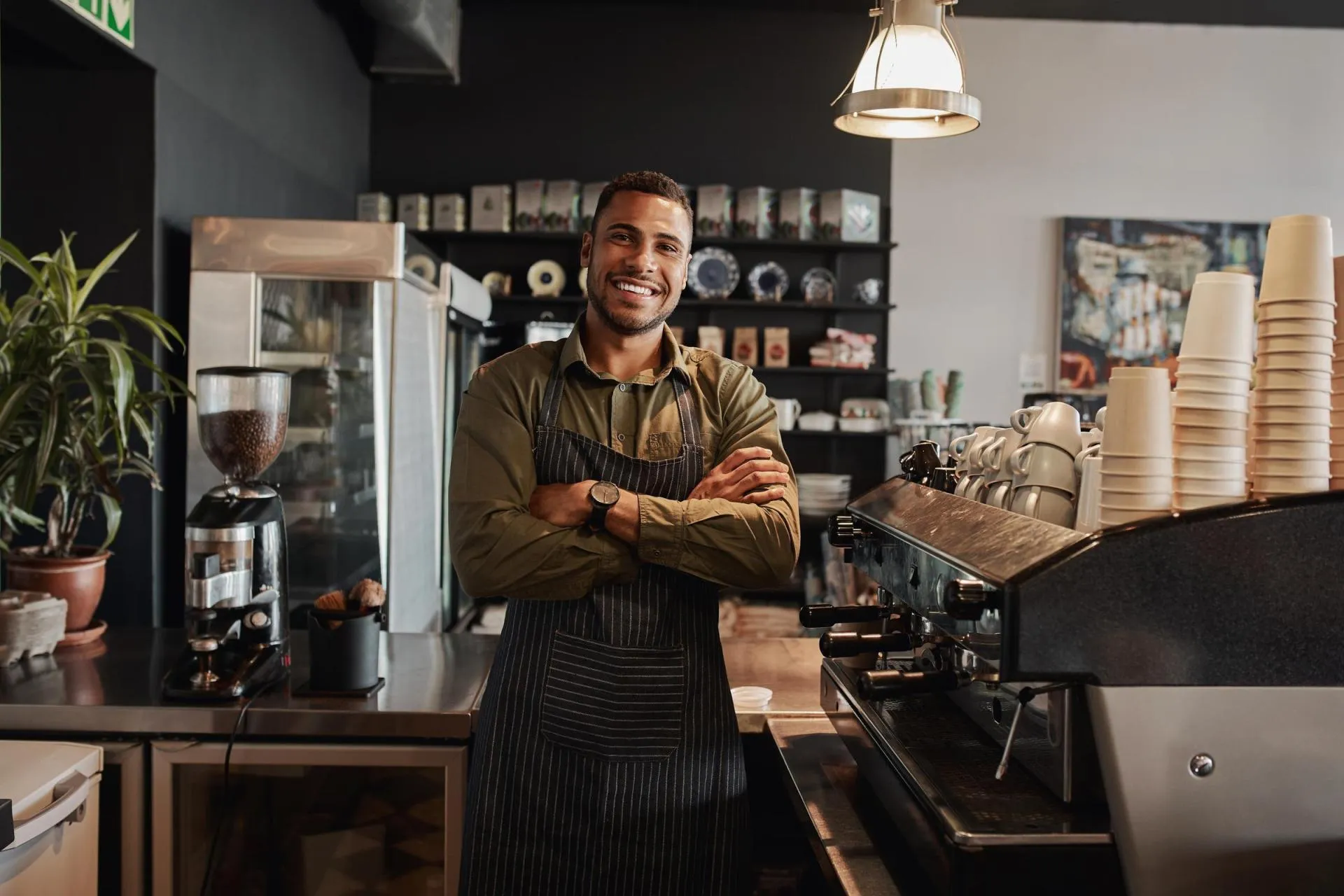 Smiling male barista standing behind the counter with crossed arms, at the coffee shop.