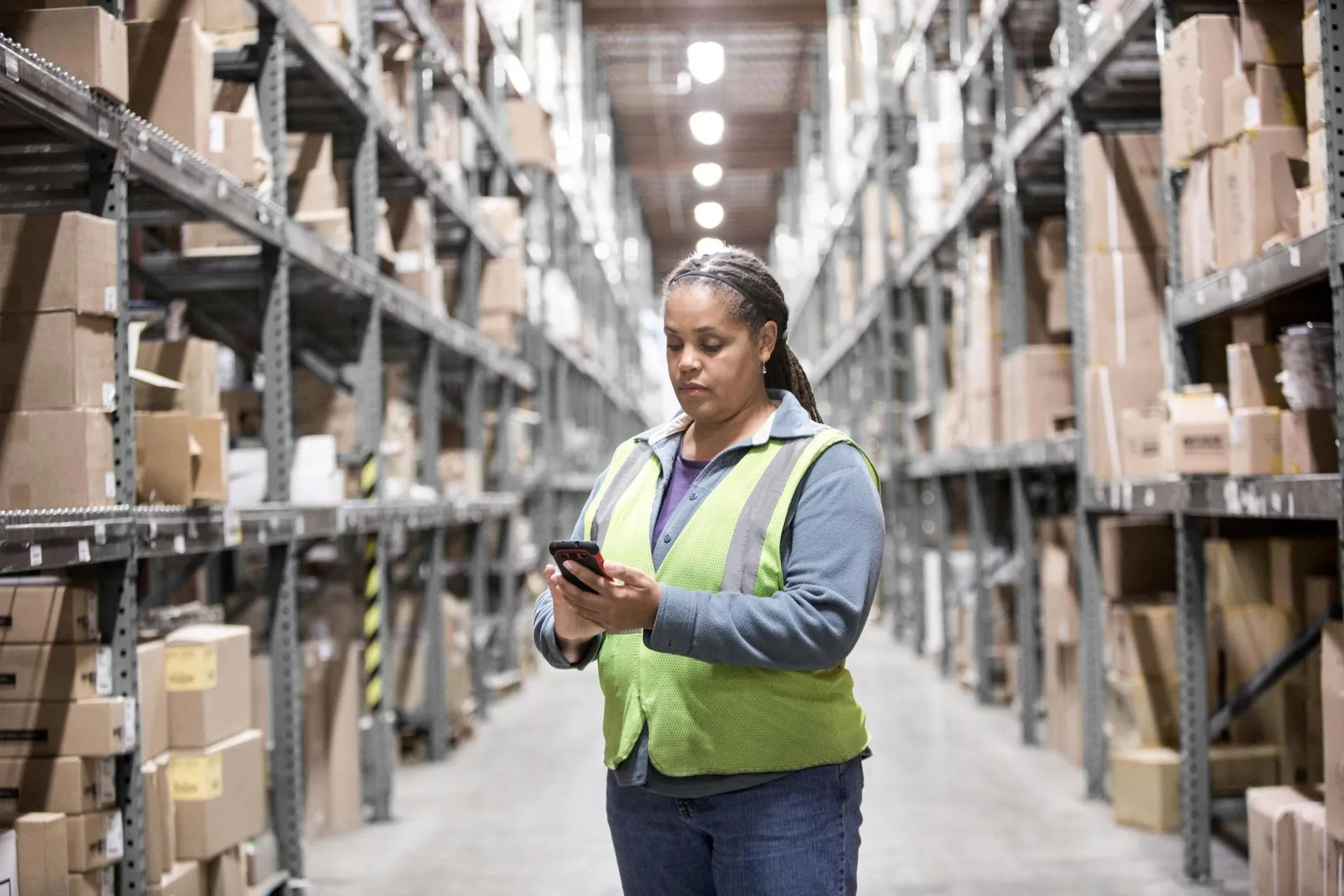 Warehouse worker wearing a high-visibility safety vest stands in the center of a storage aisle, using a handheld mobile device to manage inventory.