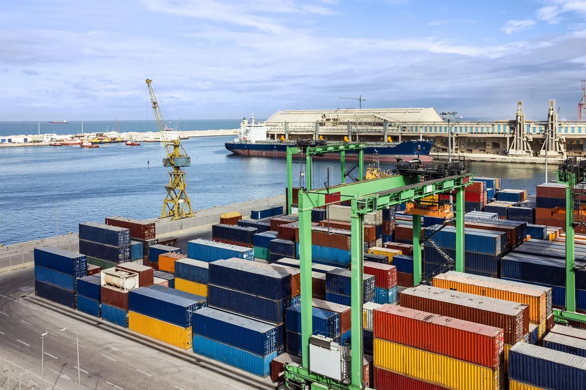 A container yard with a crane and a deep-sea carrier in the background.