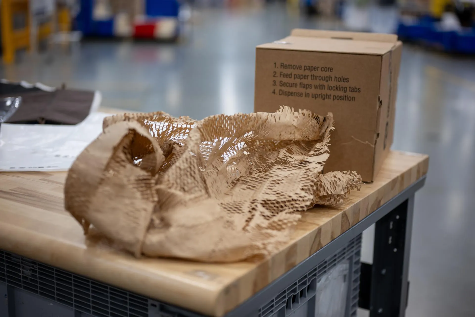 Honeycomb kraft paper packaging material spread across a wooden packing table beside a cardboard dispenser box in a warehouse.