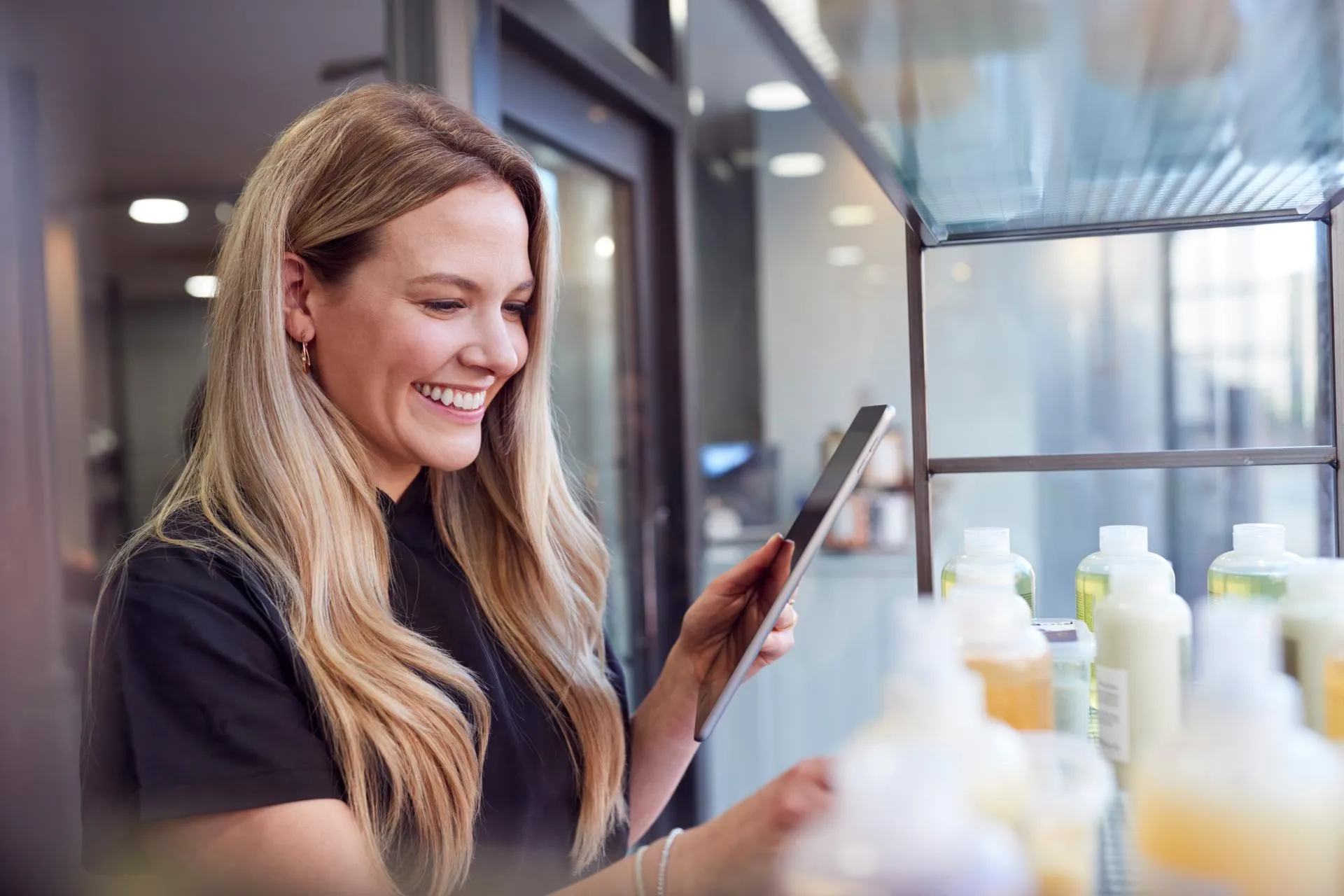 A woman with long blonde hair smiling and looking at her phone in front of a shelf with beauty bottles.