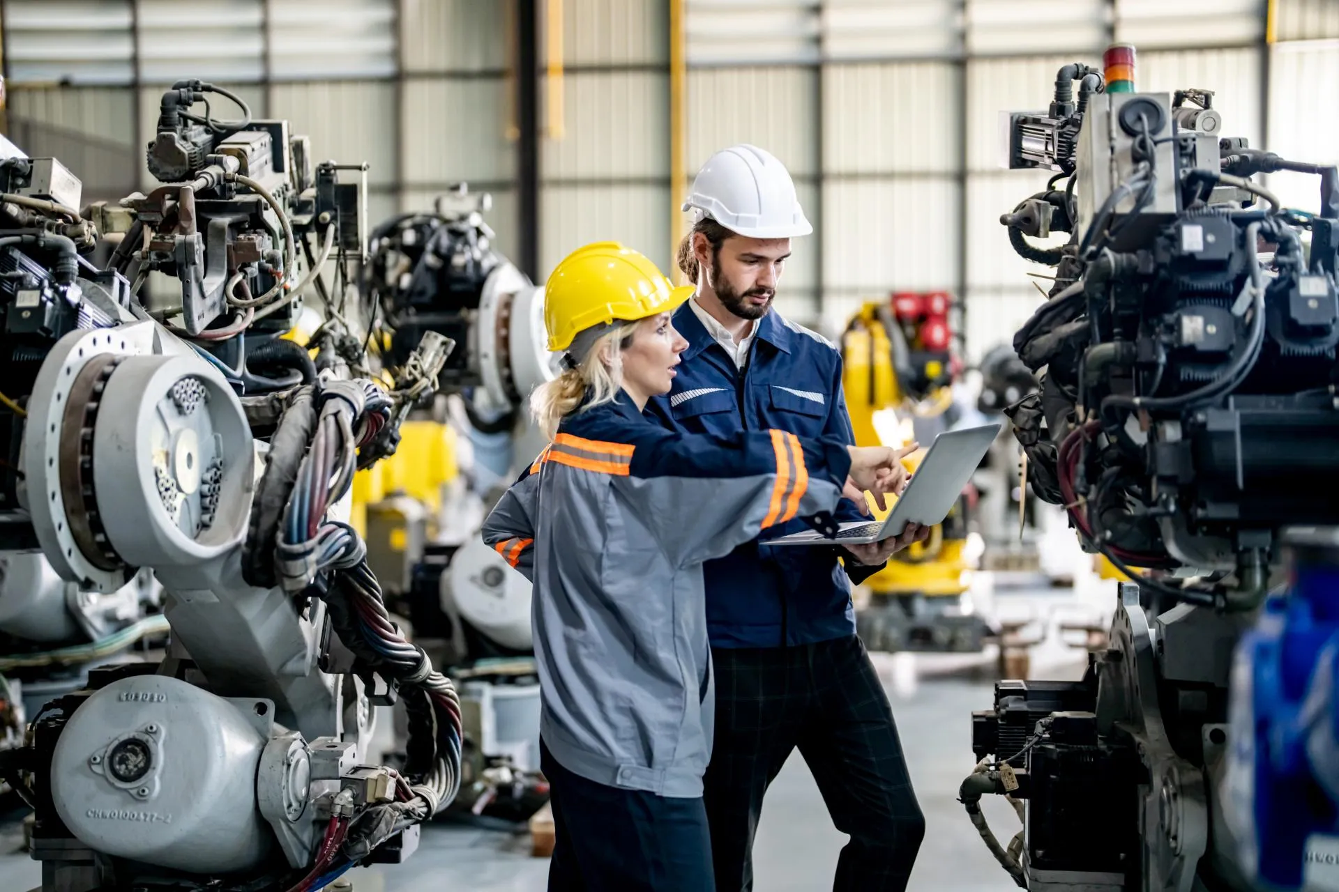 Two industrial workers in safety helmets review information on a laptop inside a factory, standing between large mechanical components and production equipment.