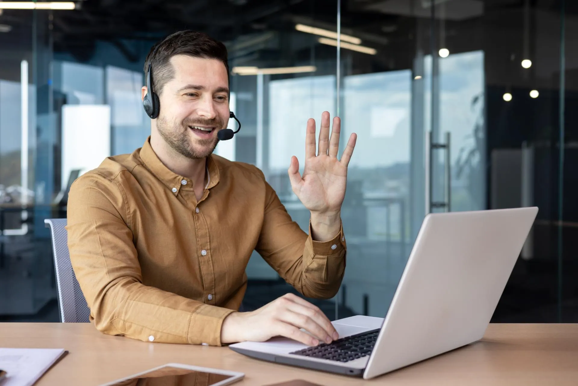 A businessman wearing a headset, smiling and waving at the camera of his laptop participates in a video call while seated at a desk in a modern office.