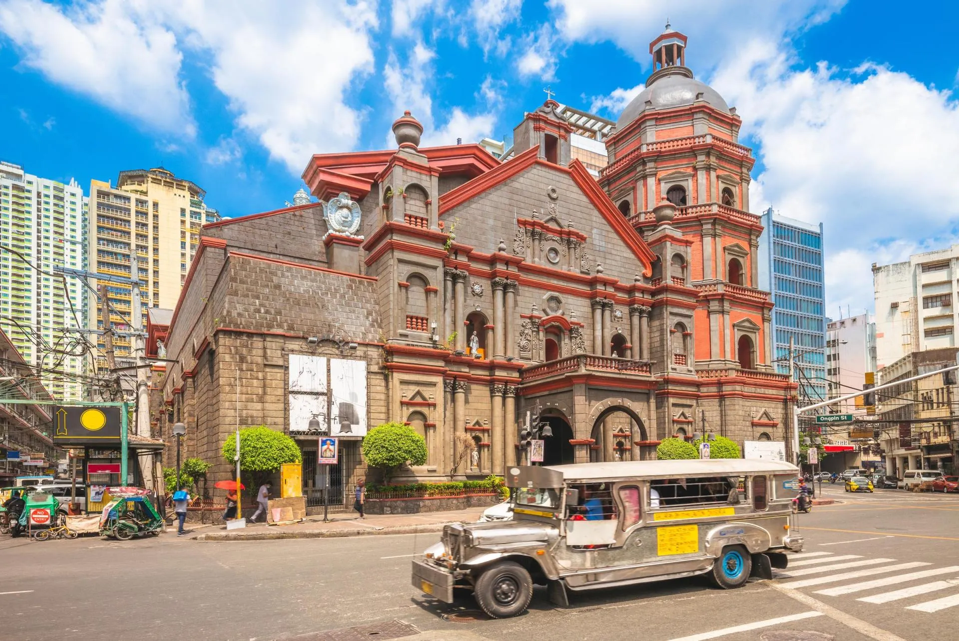 Historic church in Manila, Philippines, surrounded by modern buildings, with a traditional jeepney driving past on a busy city street under a bright blue sky.