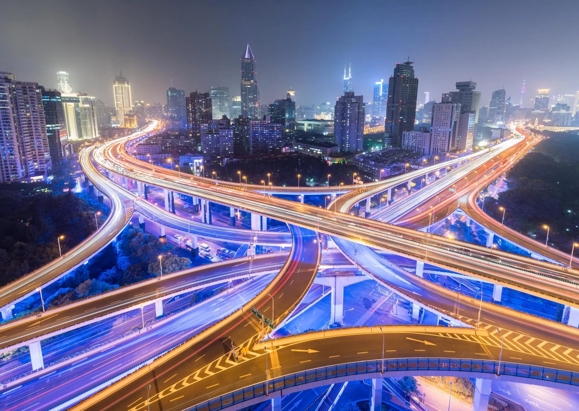 Nighttime city skyline with a multi-level highway interchange and bright long-exposure light trails. High-rise office and apartment buildings in the background.