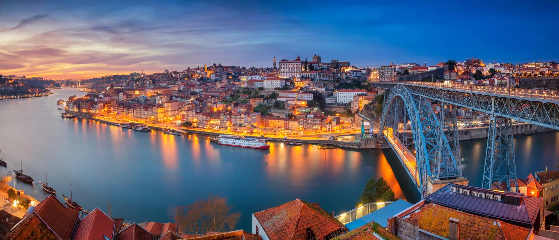 Panoramic cityscape image of Porto, Portugal, with the Luis I Bridge and the Douro River during sunset.
