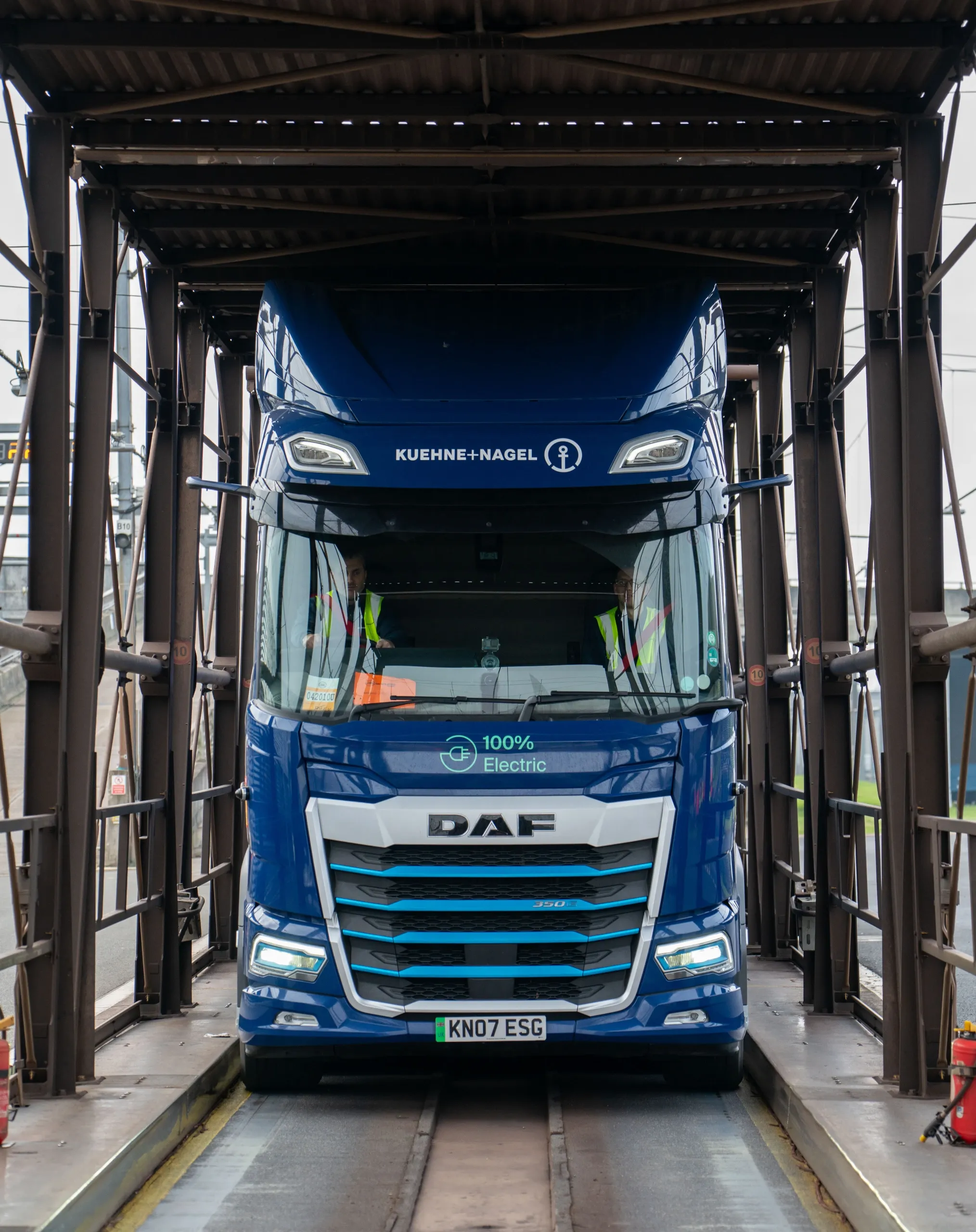Front view of a blue Kuehne+Nagel 100% electric DAF heavy‑duty truck positioned inside a loading structure.