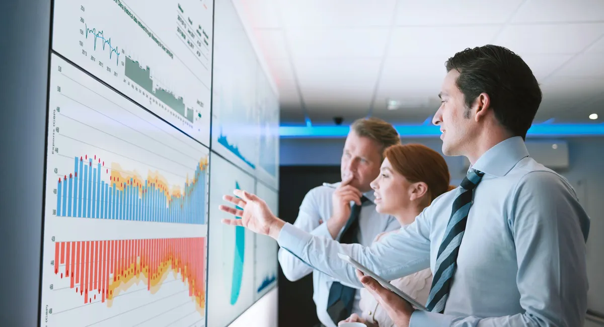 A group of people stand in front of monitor panels discussing charts.