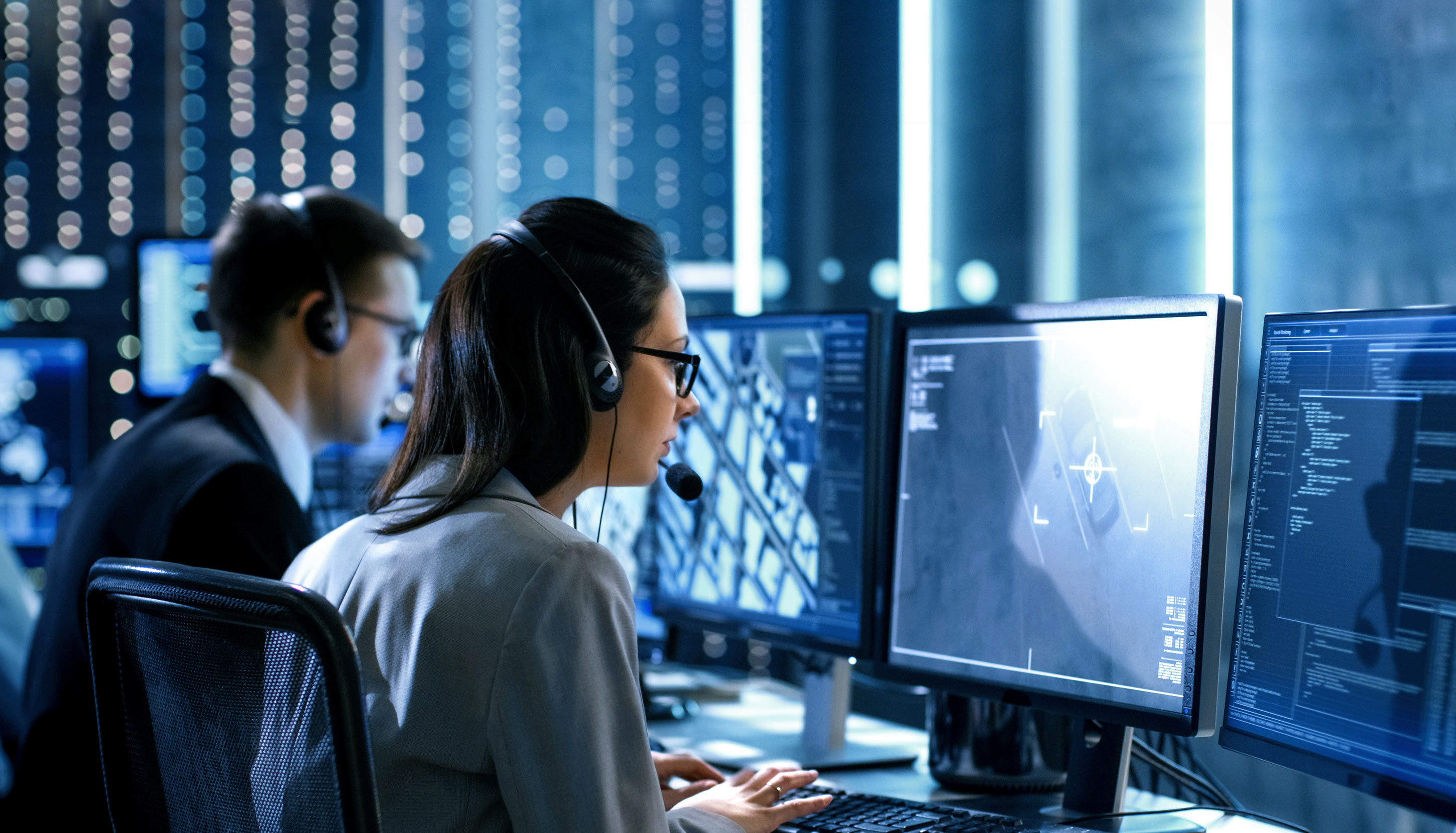 Two security workers are viewing a blue-tinted video of a car from a control center.