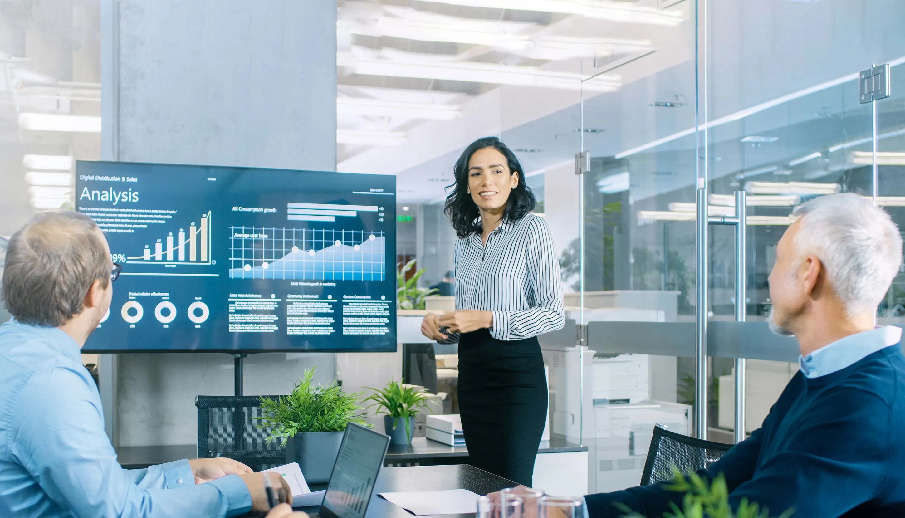 A business meeting in a modern office with three people discussing data displayed on a large screen