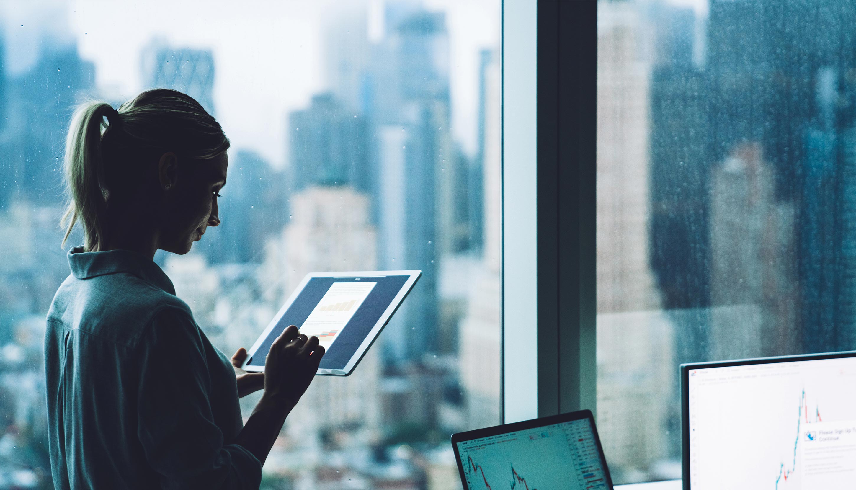 A person stands by an office window holding a tablet.