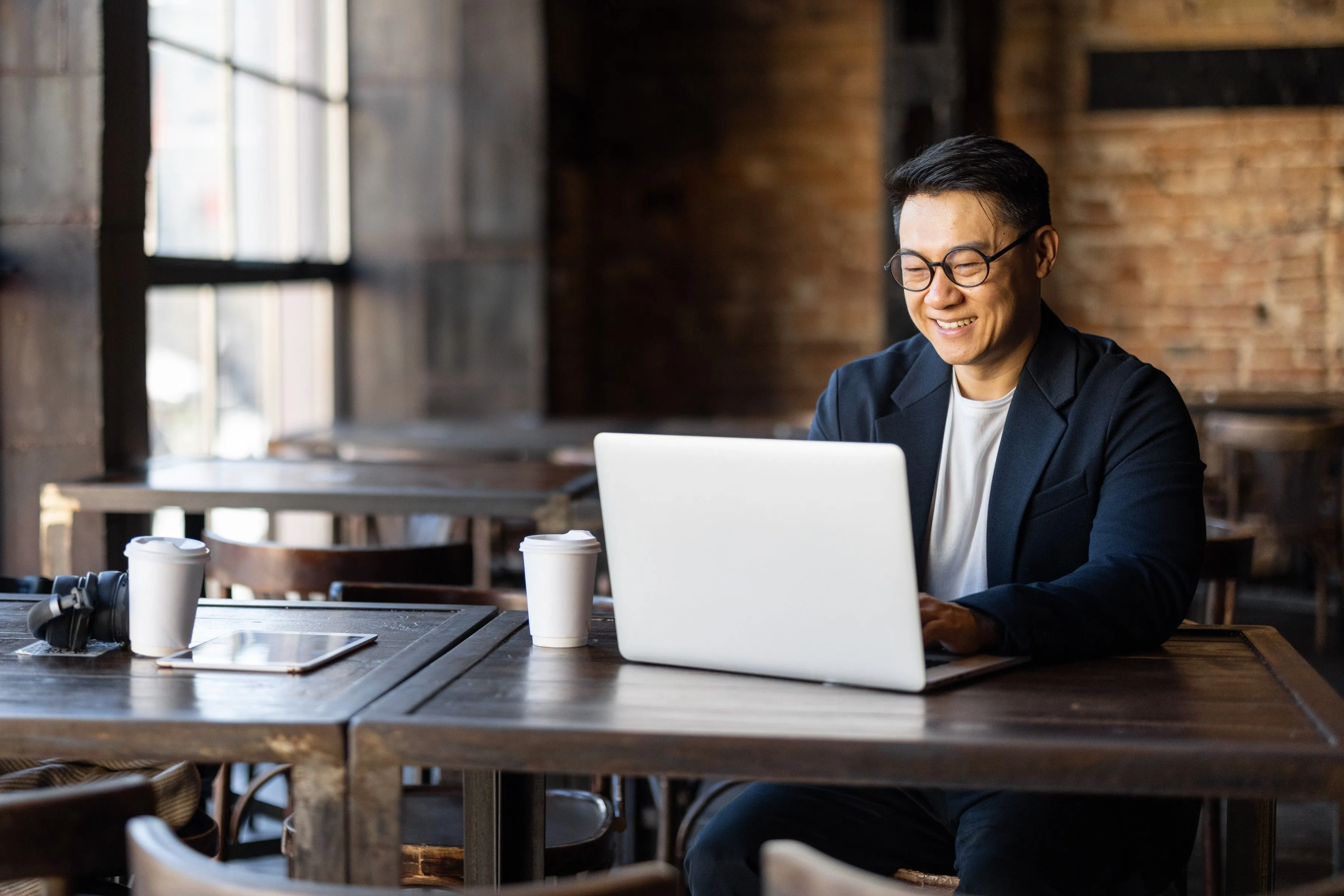 Asian businessman typing on laptop during work in cafe. Concept of remote and freelance work. Smiling adult successful man wearing suit and glasses sitting at wooden desk.