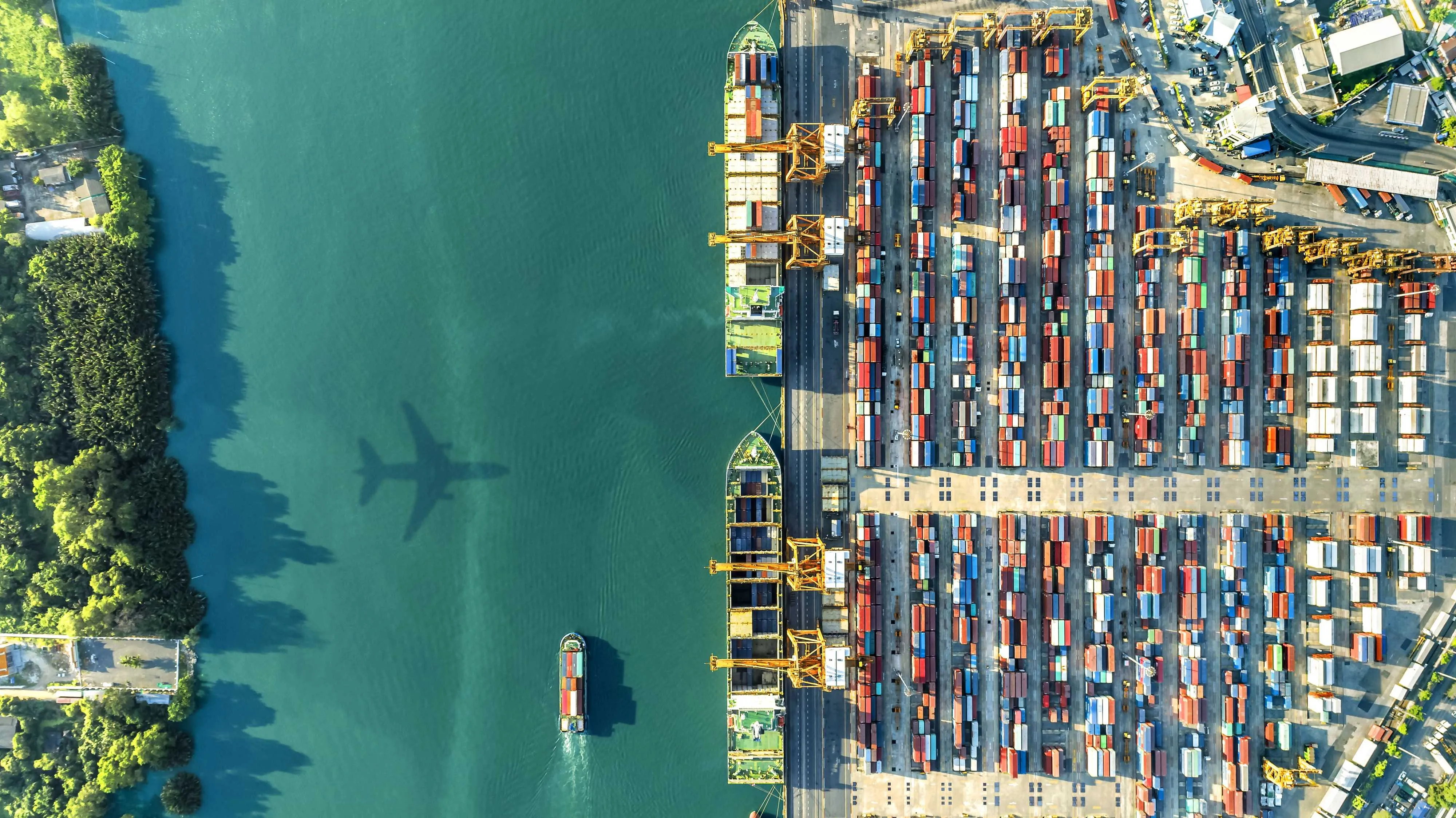 Aerial view of a container port with cargo ships docked and rows of shipping containers, with an airplane shadow visible over the water.