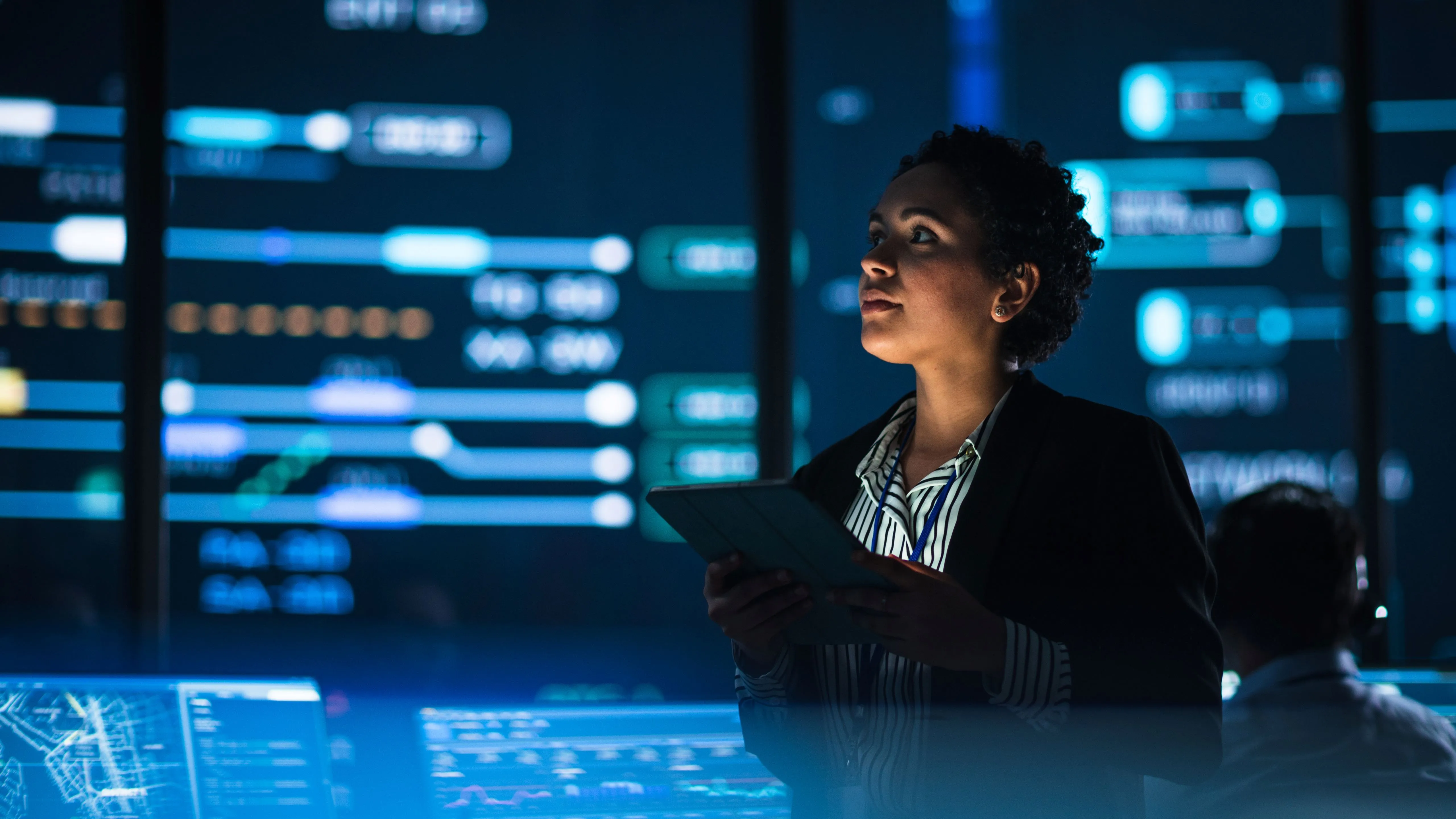Young multiethnic female government employee uses tablet computer in system control monitoring center. In the background her coworkers at their workspaces with many displays showing technical data.