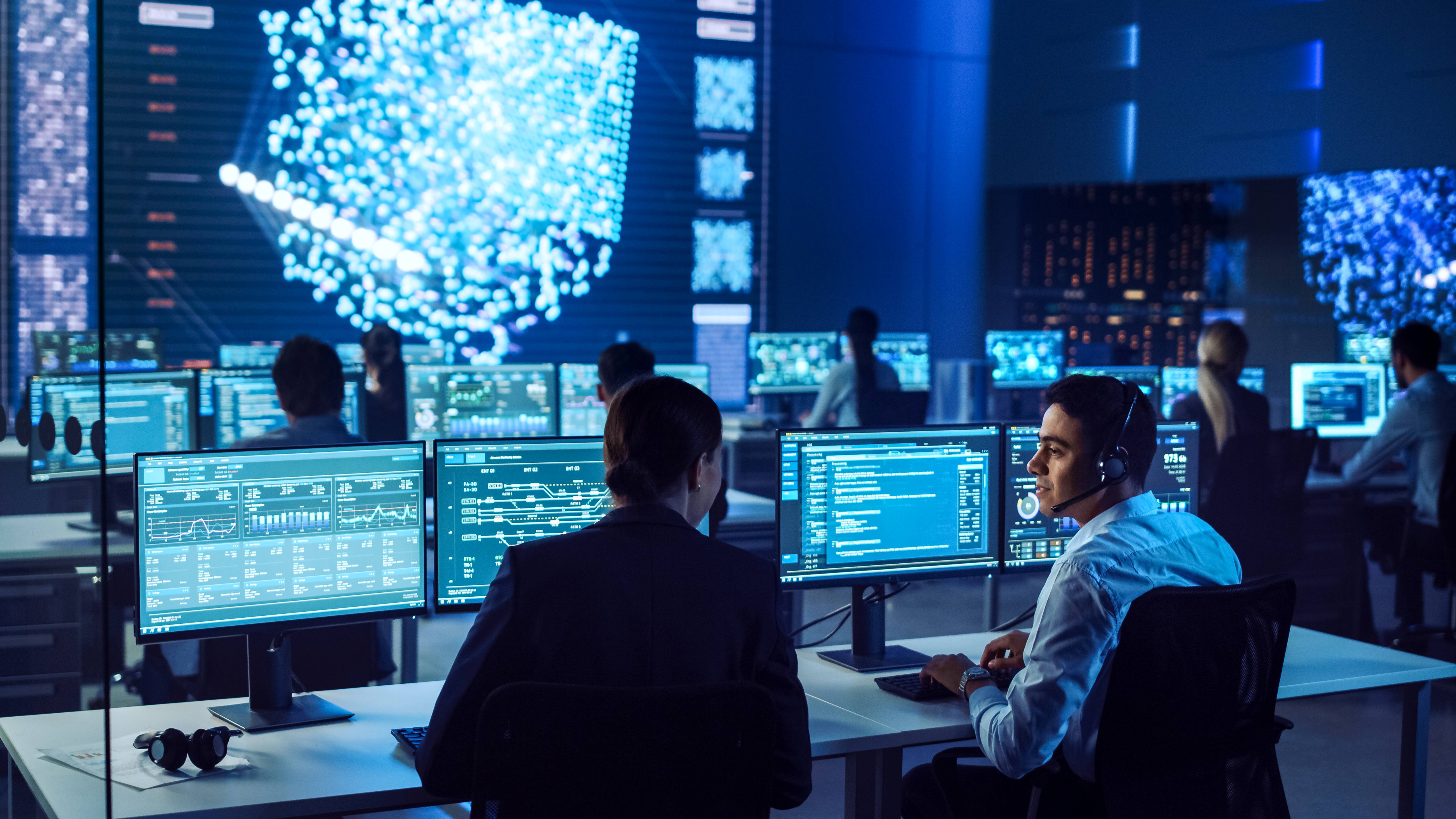 Confident female data scientist works closely to her young colleague in big control and monitoring room with neural network. Young specialists talk and discuss work in a room with other employees.