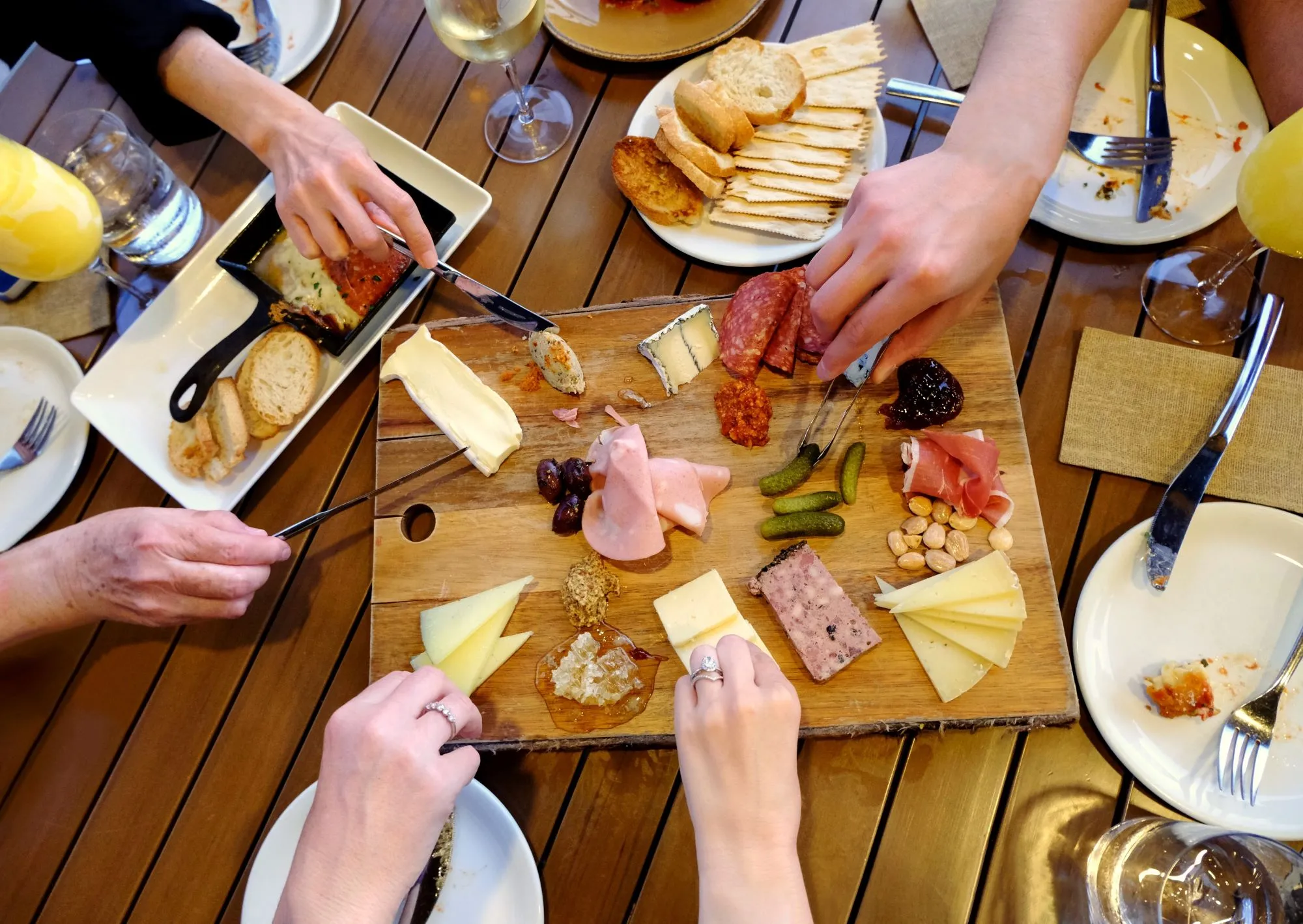 Zbliżenie na deskę z serami i wędlinami, którą dzieli się kilku przyjaciół.