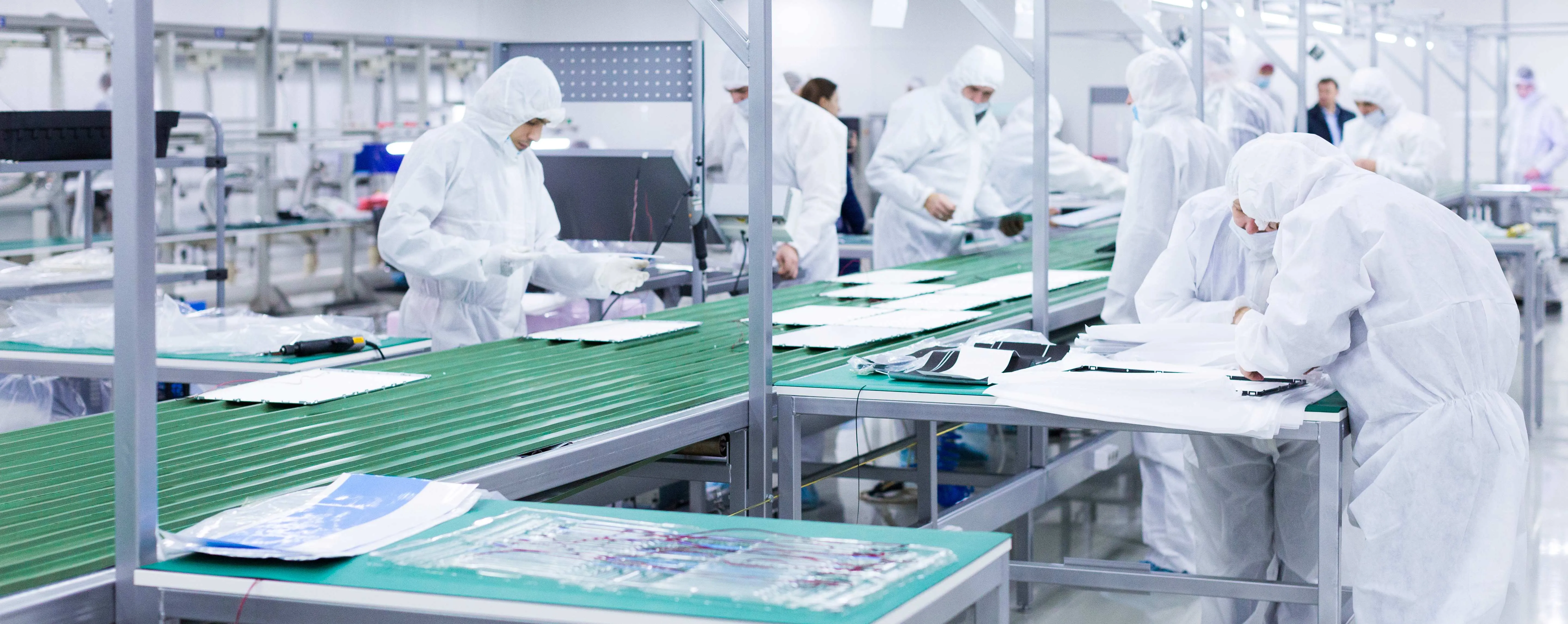 Trabalhadores de fábrica vestindo jalecos brancos e máscaras faciais, produzindo aparelhos de televisão em uma linha de montagem verde com alguns equipamentos modernos.