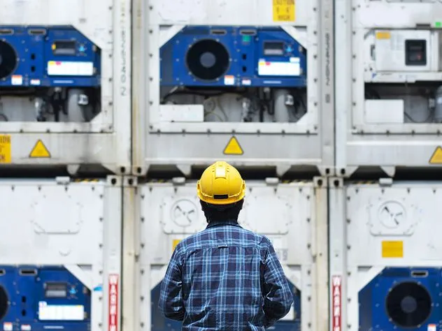 A back shot of a person in safety equipment inspecting stacked reefer containers.
