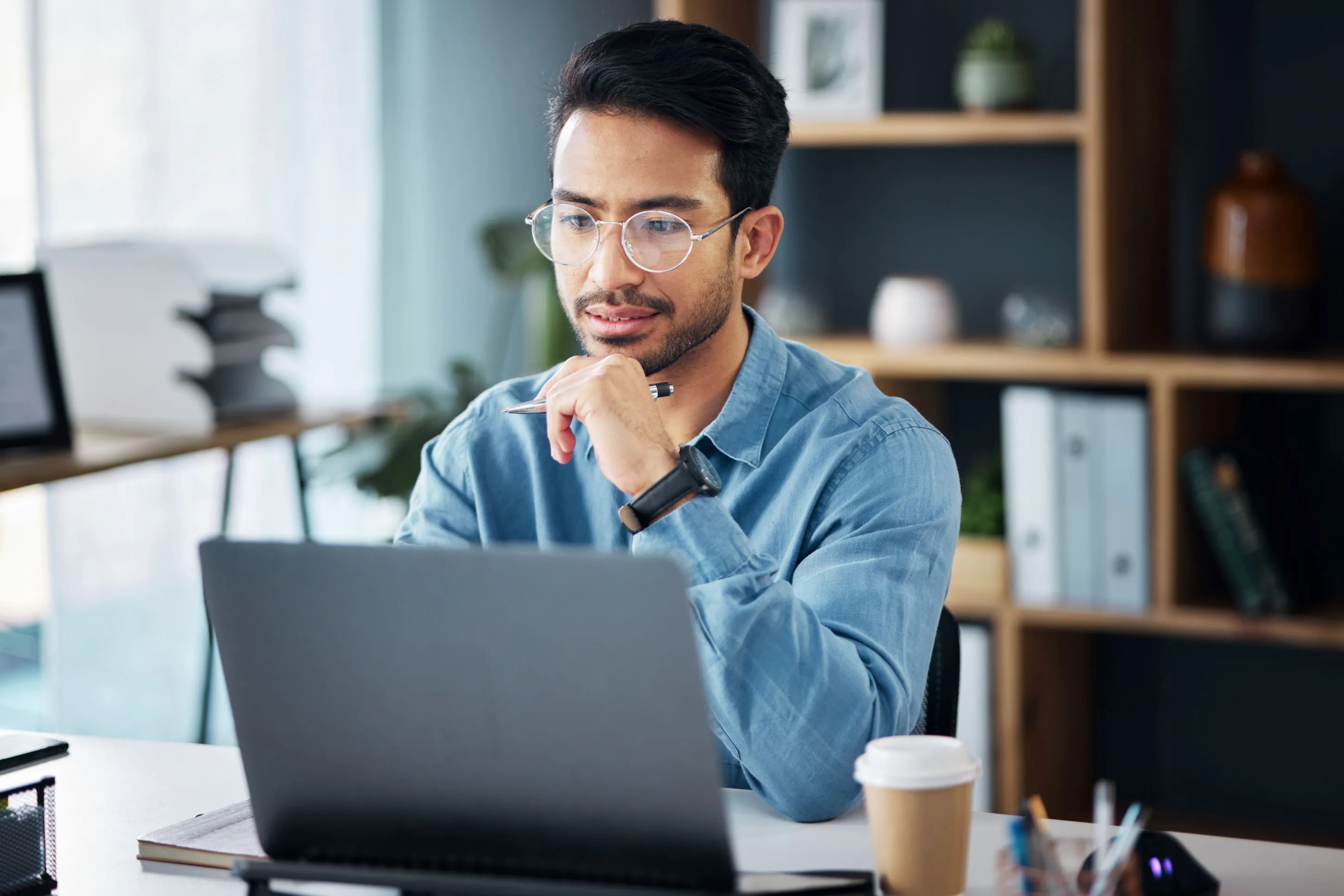 Man with glasses holding a pen while looking at his laptop.