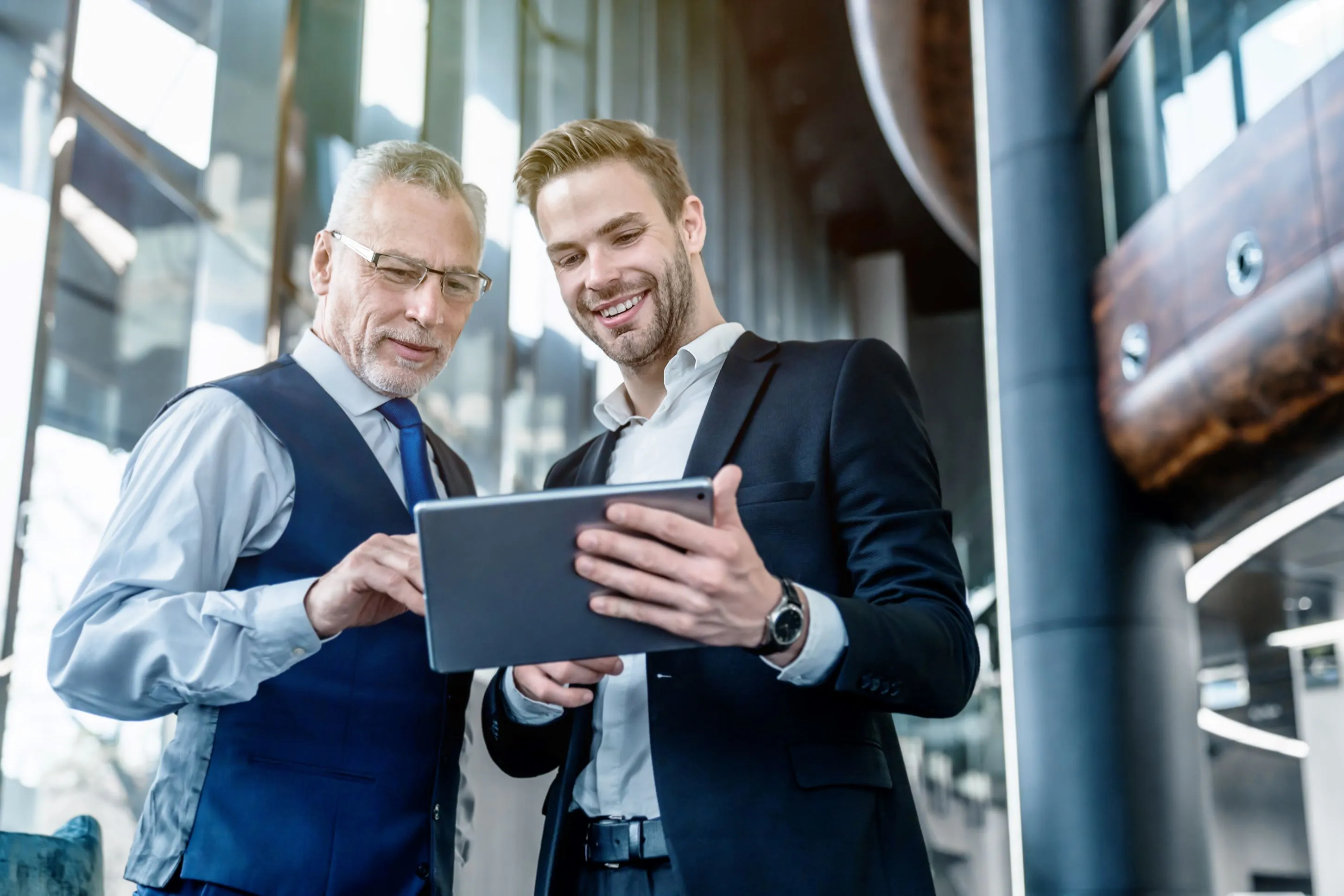 A pair of individuals stands in a tall-ceilinged office, viewing a tablet and smiling.