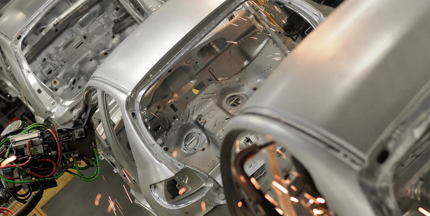 A row of grey car bodies in various stages of assembly is seen on a production line inside a brightly lit automotive factory. The image highlights the precision and scale of modern vehicle manufacturing.