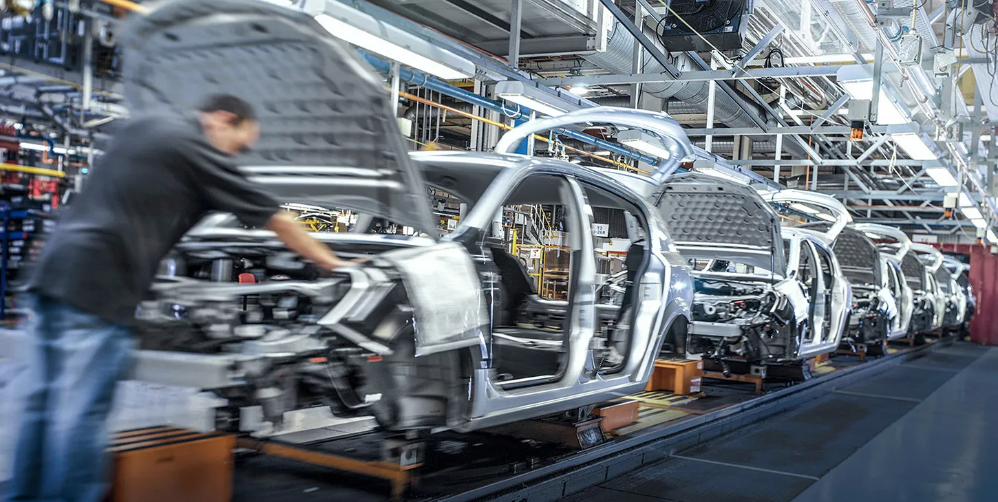 A row of grey car bodies in various stages of assembly is seen on a production line inside a brightly lit automotive factory. The image highlights the precision and scale of modern vehicle manufacturing.