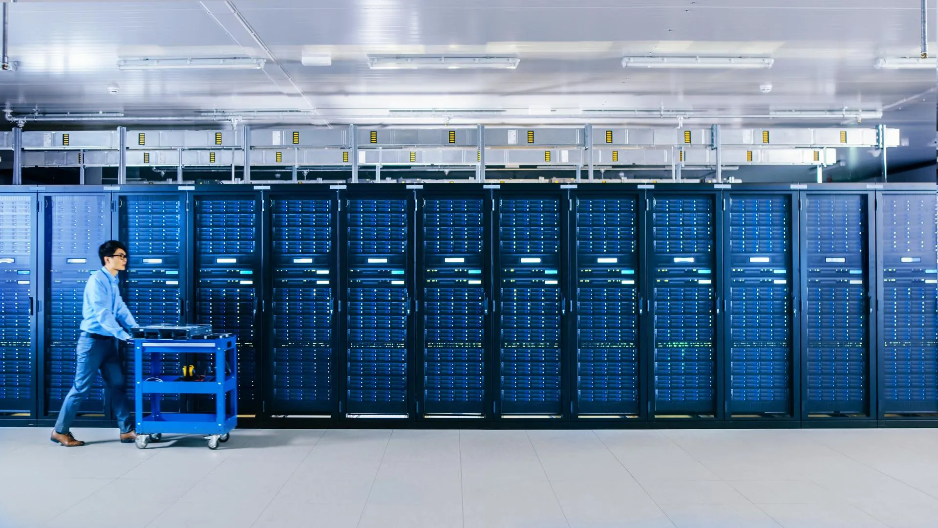 In the Modern Data Center: IT Technician Working with Server Racks, Pushes Cart Between Rows of Server Racks. On a Pushcart New Hardware for System Update. Engineer Doing Maintenance and Diagnostics.