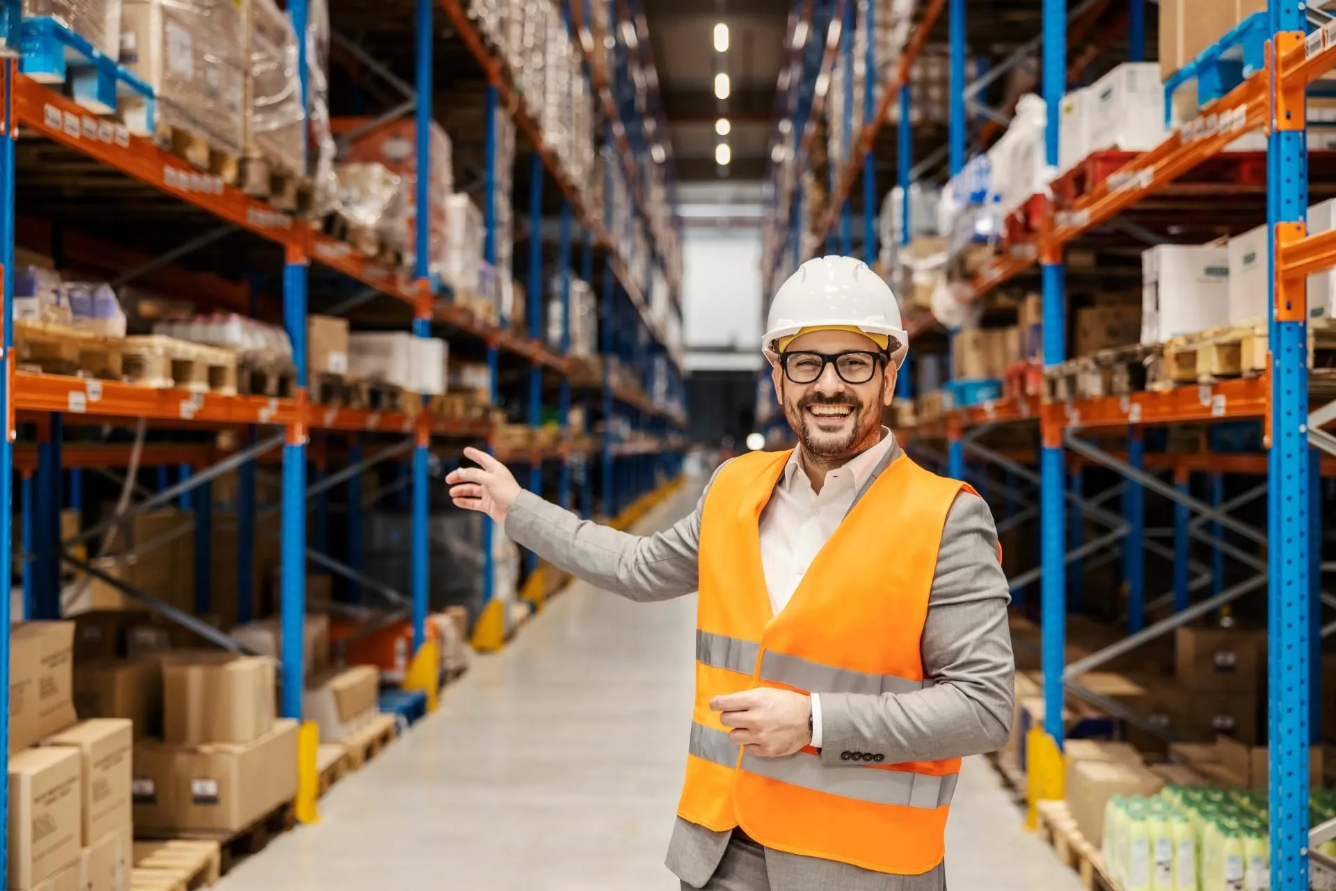 A warehouse supervisor is pointing at the commodity reserves in warehouse while smiling at the camera, there is no shortage.