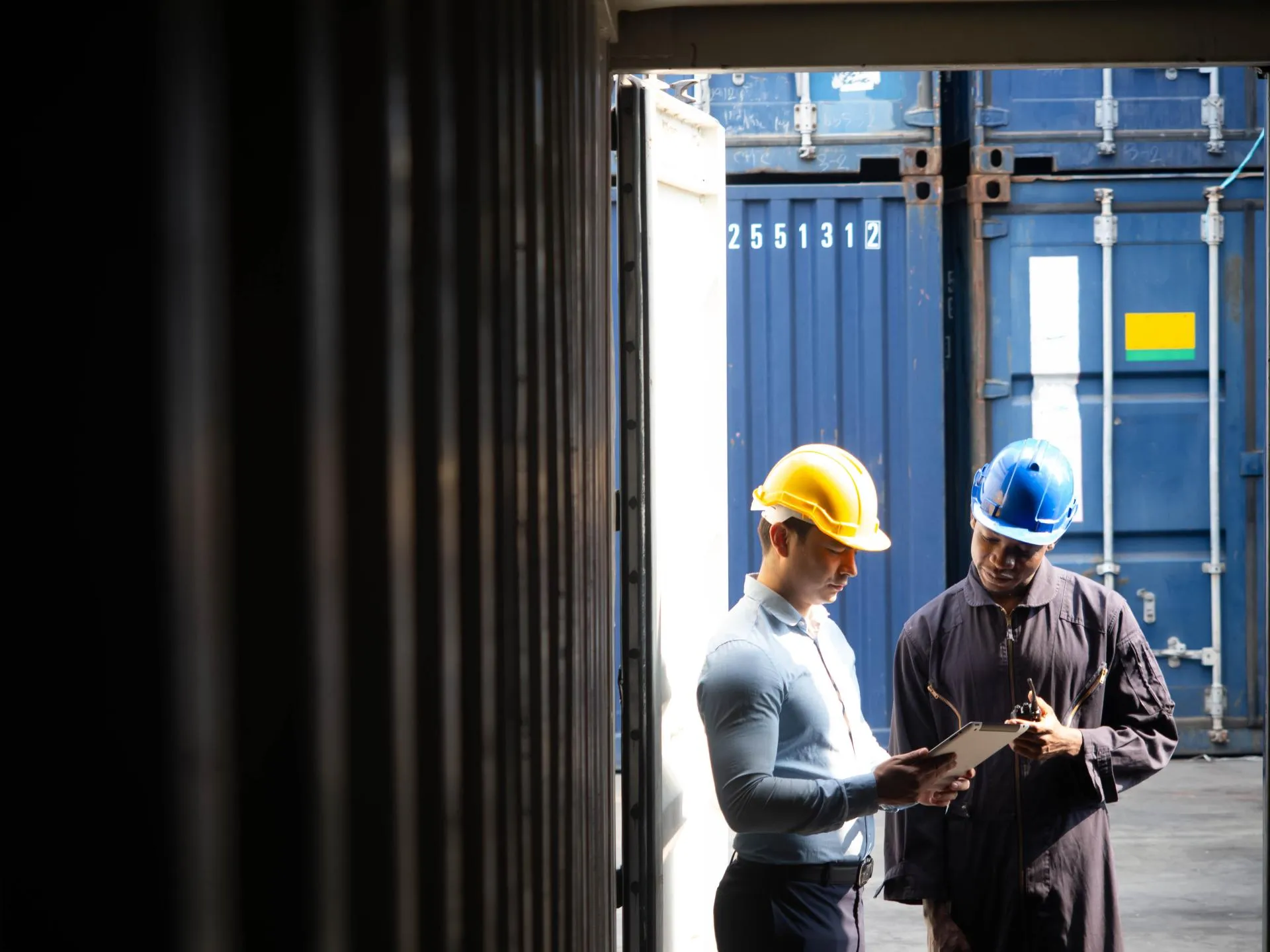 Two workers in helmets discuss near shipping containers at a warehouse