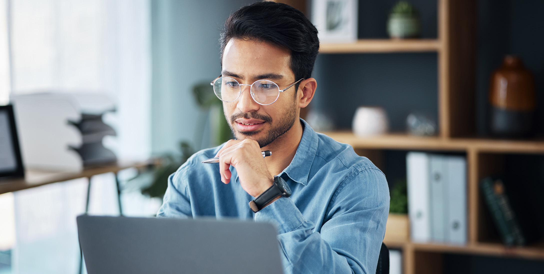 Man with glasses sitting in front of the laptop with a pen ih his hand
