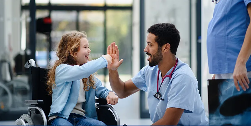 Doctor kneeling next to a girl in a wheelchair, high-five greet and smiling